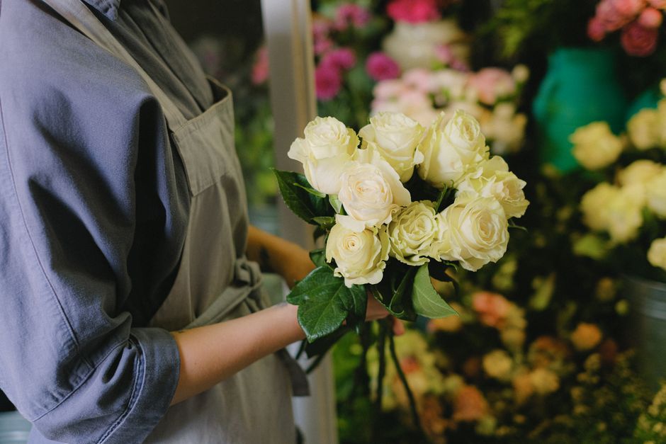 A person wearing a grey apron is holding a bouquet of fresh white roses with green leaves. The roses are arranged neatly and appear to be newly cut, with the petals soft and slightly open. The bouquet