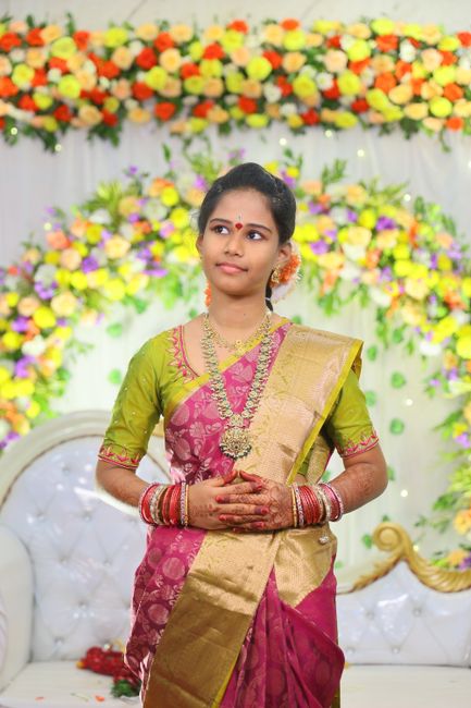 A young woman dressed in traditional Indian attire, wearing a vibrant pink and gold sari with intricate embroidery, stands in front of a festive floral backdrop at a wedding or celebration. She has ac