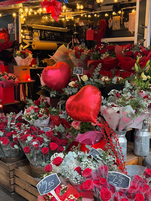 A vibrant floral display at a flower shop showcasing an array of fresh red roses, white flowers, and greenery arranged in baskets and bouquets. Two shiny red heart-shaped foil balloons are prominently