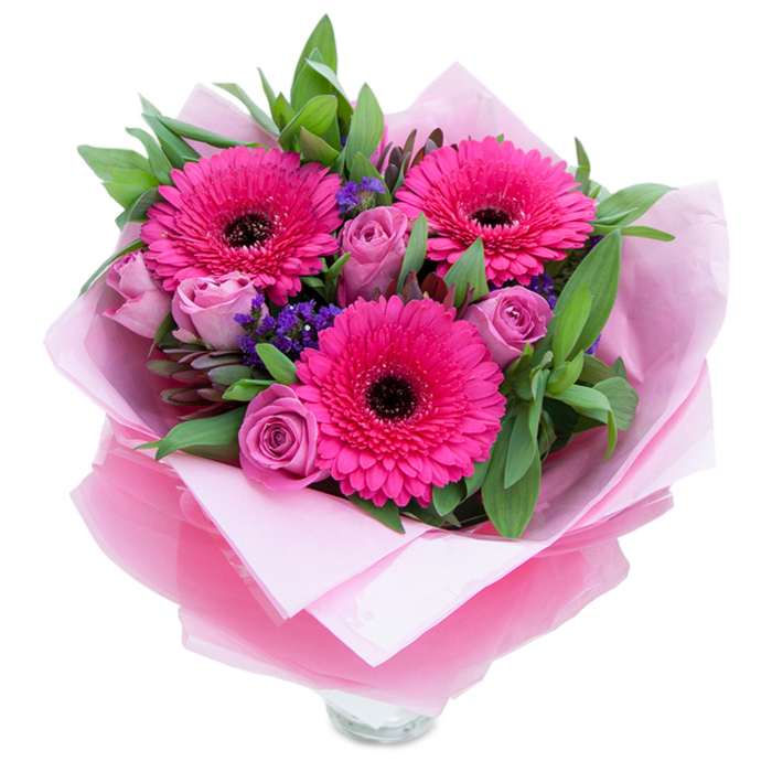 There is a graceful, garden-fresh energy to this pink bouquet, composed around three fuchsia Gerbera daisies that fan their luminous petals outward from bold, dark centres-each bloom like a little sunburst. Nestled between them are lavender-tinged roses in a dusty rose tone, their tightly furled petals forming elegant, compact cups that suggest tenderness and new beginnings. Small sprays of purple statice punctuate the arrangement with delicate pops of cool colour and a slightly papery texture that contrasts the velvety roses. The foliage is generous and green-broad leaves that recall salal or eucalyptus-providing a deep, leafy frame that lets the pinks sing without crowding them. The bouquet is lovingly wrapped: a pale pink, matte tissue cradles the stems while an outer sheet of translucent, crinkled fuchsia-pink cellophane adds a gentle shimmer and cohesive flourish; the wrapping is folded to reveal the rim of a clear glass vase below, so the flowers are ready to be displayed on a coffee table or dining sideboard. Bathed in soft studio light, the petals show subtle tonal variations and textures-the satiny gleam of rose edges, the sturdy radiance of Gerbera petals-and the scent is quietly uplifting, with rose perfume softened by a green, fresh note from the Gerberas. Thoughtfully balanced and joyful, this arrangement is perfect for marking birthdays, Mother's Day, anniversaries or for sending a cheerful hello to friends and family across Bromley Common and nearby Sundridge Park.